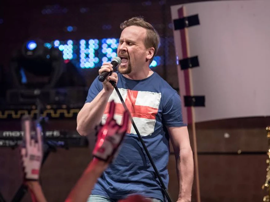 Man står och sjunger i mikrofon på en scen med ljus i bakgrunden. Konsertbild med blå T-shirt och publik i förgrunden.