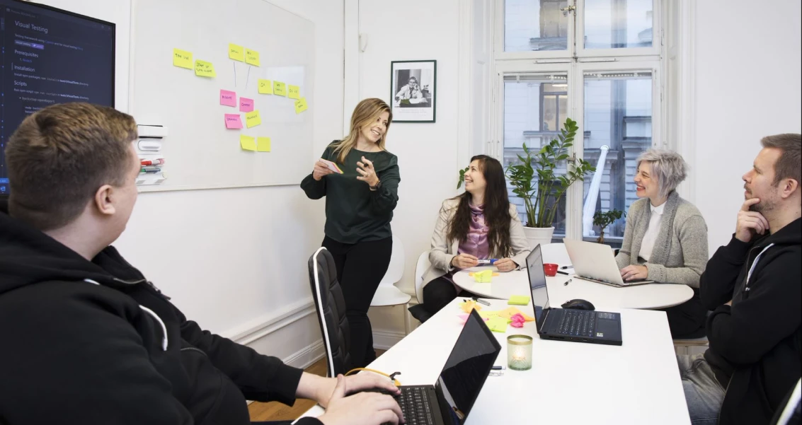 Personer i ett möte i ett kontorsrum, använder laptops och brainstormar vid whiteboard med färgglada post-it-lappar. 