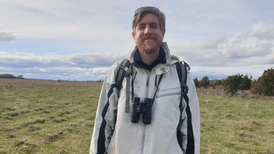 A person standing in an open field wearing a white Helly Hansen jacket, with binoculars around their  neck, under a cloudy sky