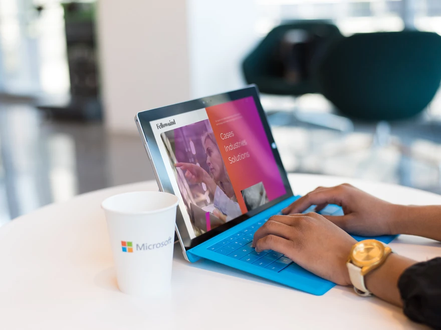 Person som skriver på en surfplatta vid ett bord med en Microsoft-kopp, i en ljus kontorsmiljö.