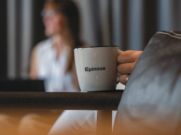 En kaffemugg i fokus i bild, med person i bakgrunden sittandes i ett rum