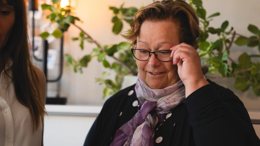 N&auml;rbild av en person i svart kl&auml;dsel med lila scarf och glas&ouml;gon, sittande inomhus med gr&ouml;na v&auml;xter i bakgrunden.