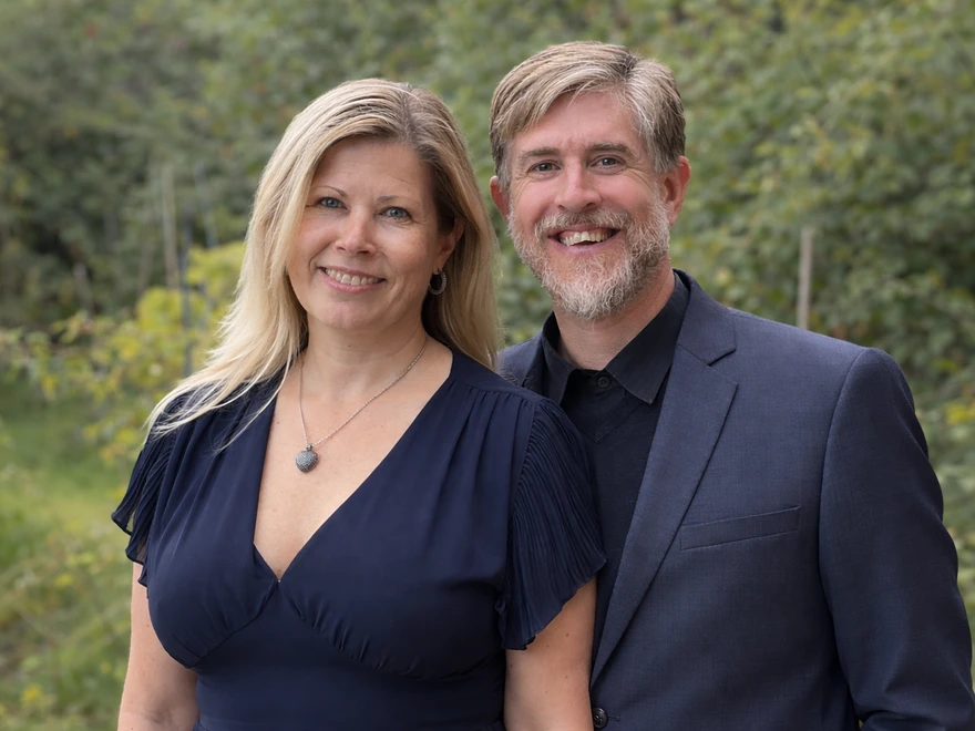 Portrait of a couple standing outdoors, with the woman in a navy dress and necklace and the man in a dark suit and black shirt