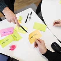 Personer i möte diskuterar projektledning med färgglada post-it-lappar märkta med termer som "Web Pack," "Azure" och "Hosting" på skrivbordet. En laptop syns i hörnet
