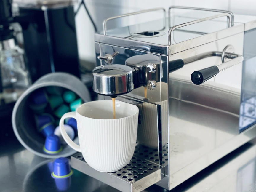 Espressomaskin i rostfritt stål brygger kaffe i en vit mugg, med kaffekapslar på sidan i en behållare