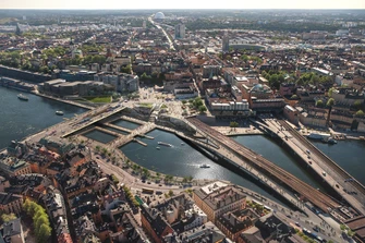 Luftvy över centrala Stockholm med Slussen och djurgårdsfärjan i förgrunden, moderna byggnader och Gamla Stan synliga i bakgrunden
