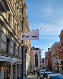 Flagga med texten "Epinova" på en byggnad i en stadsgata med klassisk arkitektur, klar himmel i bakgrunden