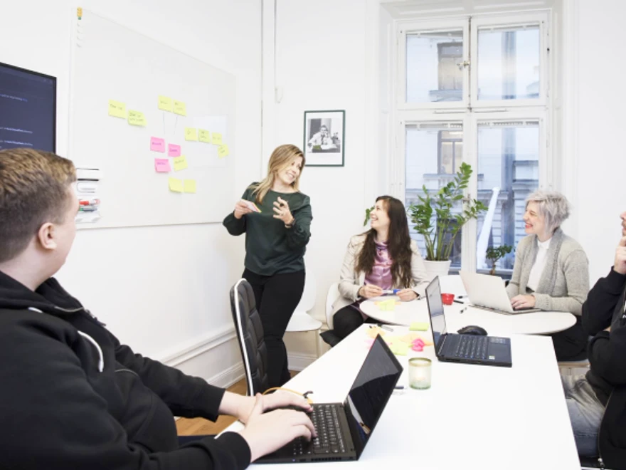 Personer i ett möte i konferensrum diskuterar framför en whiteboard med klisterlappar. 